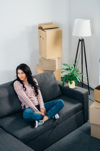 cheerful latin woman sitting on sofa with crossed legs in new home - Photo, Image