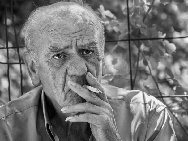 Крупный план портрета серьезного старого грека, пенсионера, который курит сигарету с улыбкой, в черно-белом
 - Фото, изображение
