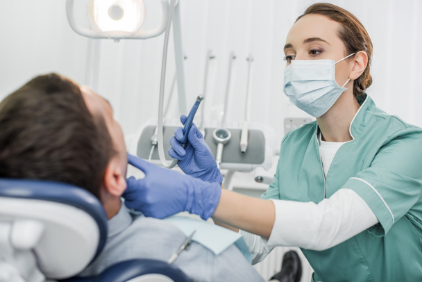 female dentist in mask holding dental instrument and touching face of patient in dental clinic - Foto, Bild