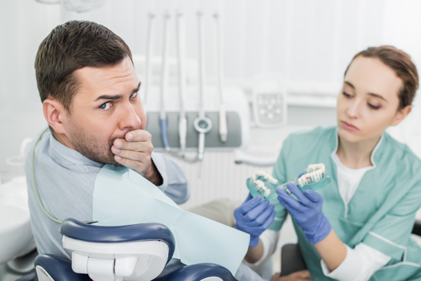 επιλεκτική εστίαση των όμορφος άντρας που καλύπτουν το στόμα με το χέρι, κοντά στο όμορφο οδοντίατρος κρατώντας τα δόντια μοντέλο  - Φωτογραφία, εικόνα