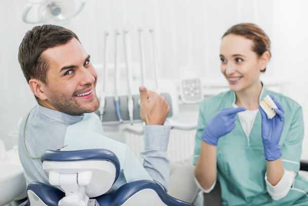 selective focus of handsome man showing thumb up near beautiful dentist holding teeth model  - Photo, Image