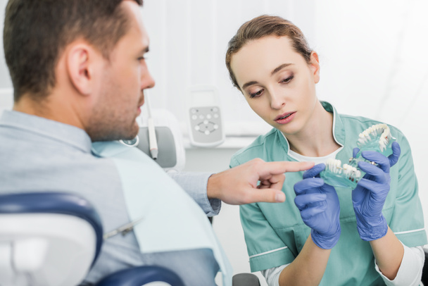 селективный фокус человека, указывающего пальцем на модель зубов рядом с красивым стоматологом
  - Фото, изображение