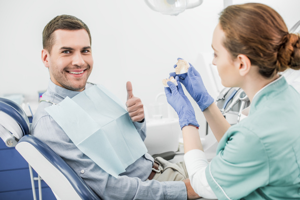 selective focus of happy man showing thumb up near female dentist holding teeth model  - Fotografie, Obrázek