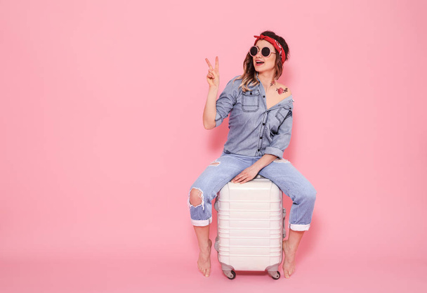 Горизонтальний портрет стильної дівчини в сонцезахисних окулярах і джинсовий одяг з татуюванням на її тілі, що сидить на білій валізі на рожевому тлі, концепція способу життя і подорожей
 - Фото, зображення