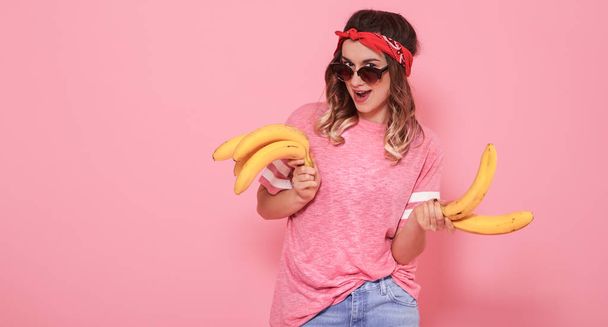 Retrato horizontal de menina hipster em roupas elegantes com óculos de sol, com bananas em suas mãos, em um fundo rosa, conceito de estilo de vida
 - Foto, Imagem