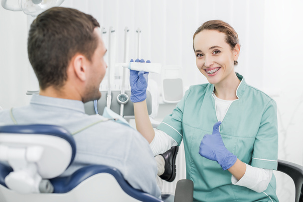 Selektywny fokus wesoły kobieta dentysty gospodarstwa pasty do zębów i pokazuje kciuk w pobliżu pacjenta - Zdjęcie, obraz