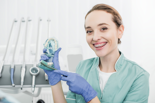 hermoso dentista con frenos en los dientes sosteniendo los dientes modelo y mostrando cómo cepillarse los dientes con cepillo de dientes
  - Foto, Imagen