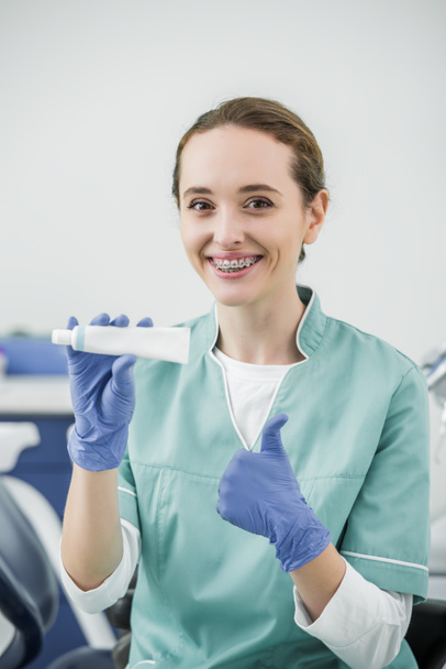 cheerful female dentist with braces on teeth holding toothpaste and showing thumb up - Φωτογραφία, εικόνα