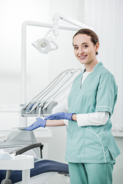 attractive dentist with braces on teeth smiling while gesturing in dental clinic  - Foto, Bild