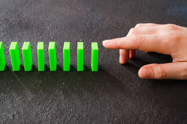 A kéz készen áll a dominószeletre, hogy láncreakciót okozzon. Zöld dominók sora sötét háttérrel - Fotó, kép