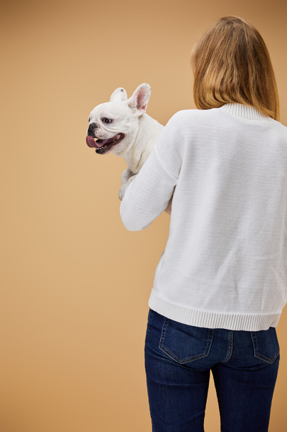 Frau in weißem Pullover und blauer Jeans mit französischer Bulldogge auf beigem Hintergrund - Foto, Bild