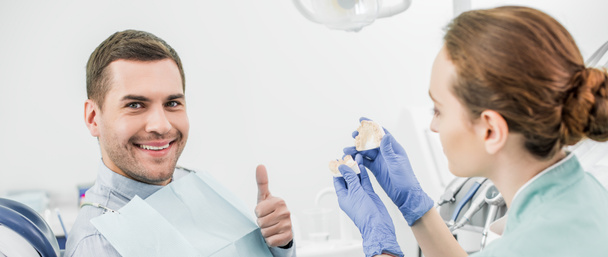 enfoque selectivo del hombre guapo mostrando el pulgar hacia arriba cerca del dentista femenino sosteniendo el modelo de dientes
  - Foto, Imagen