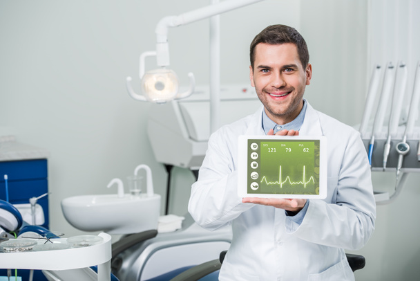 cheerful dentist holding digital tablet with medical app on screen in dental clinic  - Photo, Image