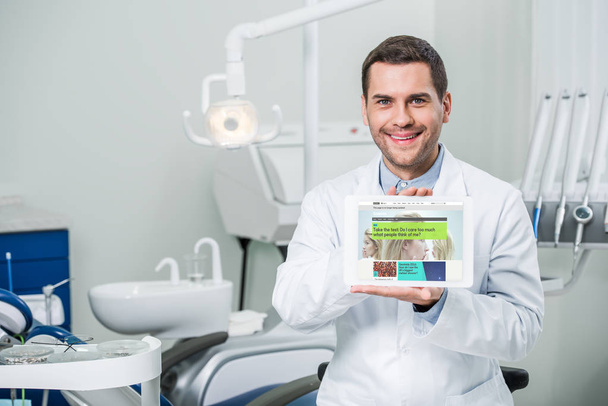cheerful dentist holding digital tablet with bbc app on screen in dental clinic  - Photo, image