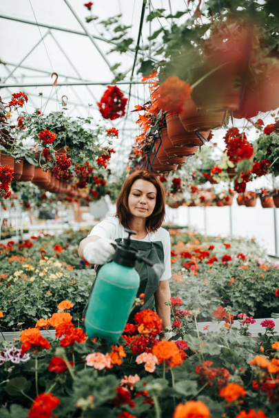 Redhead young woman working in greenhouse and enjoying in beautiful flowers. Female worker working at garden center. - Фото, изображение