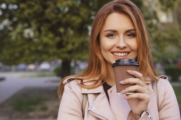Red haired sexy plus size woman walking on a park. Cold weather, she wears leather jacket. She drinks coffee from "to go" cup. Stunning sunset on a background. Copy space - 写真・画像