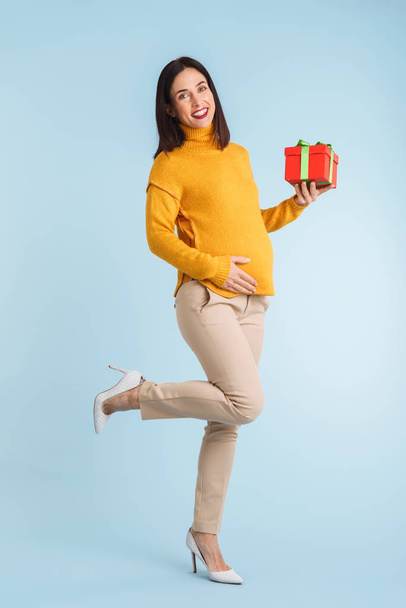 Photo of a young pregnant woman isolated over blue wall background holding gift box. - Foto, Imagen