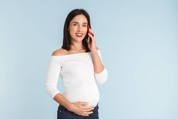 Photo of a young pregnant woman isolated over blue wall background talking by mobile phone. - Foto, Imagem