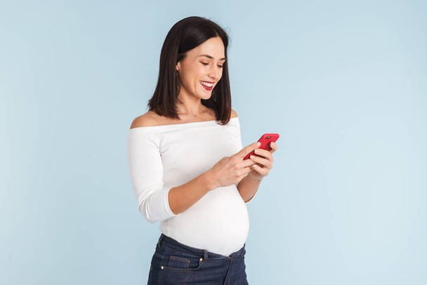 Photo of a young pregnant woman isolated over blue wall background using mobile phone. - Foto, Bild
