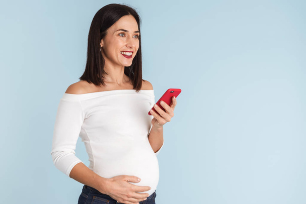 Photo of a young pregnant woman isolated over blue wall background using mobile phone. - Photo, image