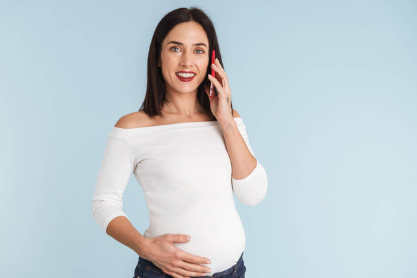 Photo of a young pregnant woman isolated over blue wall background talking by mobile phone. - Фото, зображення