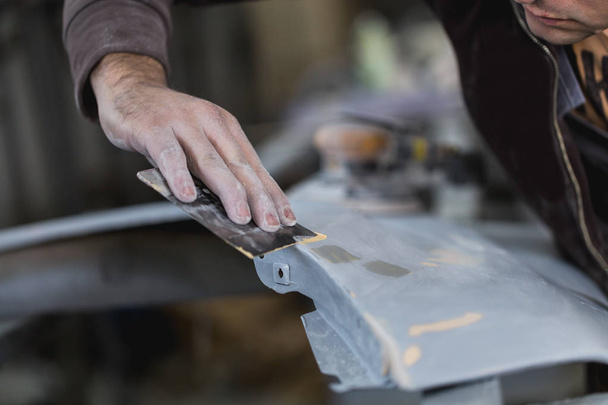 Car detailing - Man with sandpaper in auto repair shop sanding polishing and preparing car parts for painting. Selective focus on man's hand. - Photo, image