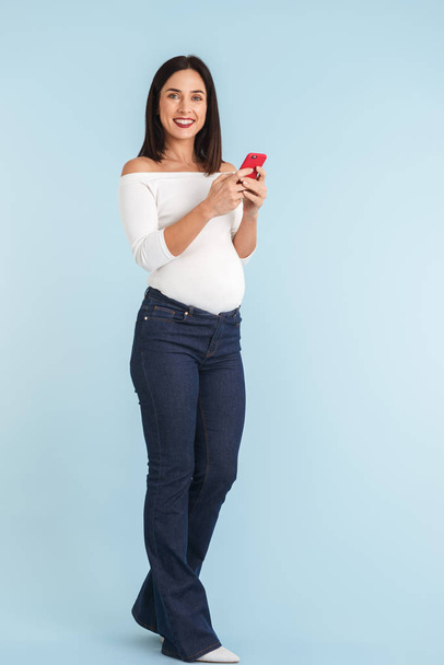 Photo of a young pregnant woman isolated over blue wall background using mobile phone. - Фото, изображение