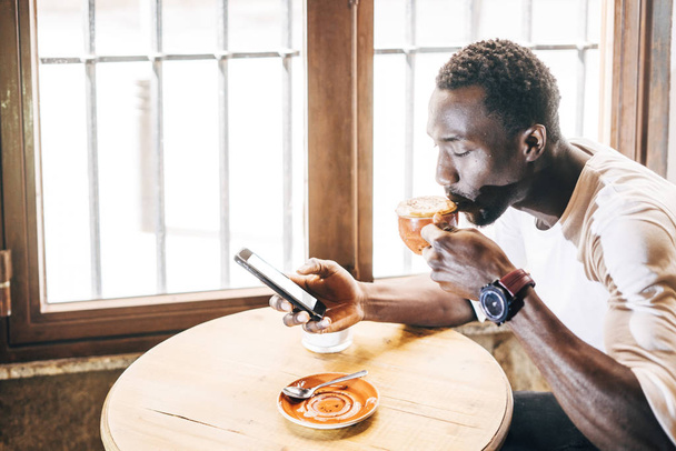 Αφρικανική άνθρωπος συμβουλεύεται το κινητό τηλέφωνο ενώ έχοντας ένα καφέ. - Φωτογραφία, εικόνα