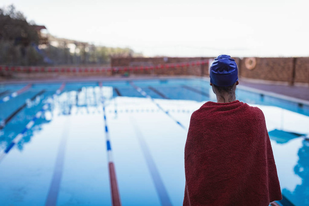 Вид сзади на молодую пловчиху, завернутую в полотенце, стоящую у бассейна
 - Фото, изображение