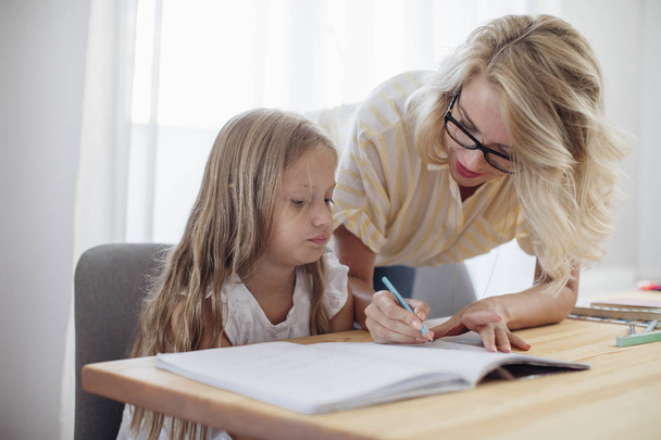 Beautiful blonde Caucasian woman helping to her cute daughter to study at home. - Fotó, kép