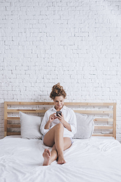 Pretty smiling Caucasian woman having a good time using her mobile phone while waking up in her bed. - Foto, Bild