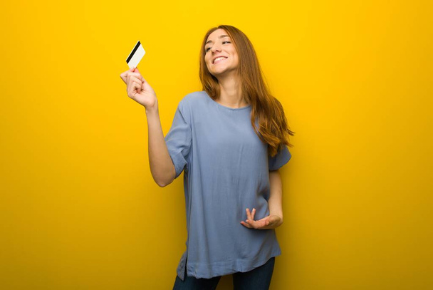 Jonge roodharige meisje over gele muur achtergrond een credit card bedrijf en denken - Foto, afbeelding