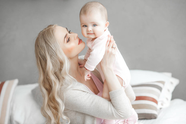 Young mother taking care of her little baby girl. Beautiful mom and her daughter indoors in the bedroom. Loving family. Attractive mum holding her child. - Zdjęcie, obraz