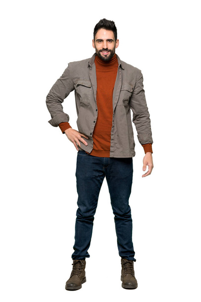 Full-length schot van knappe man met baard poseren met de armen bij de hip en glimlachend op geïsoleerde witte achtergrond - Foto, afbeelding