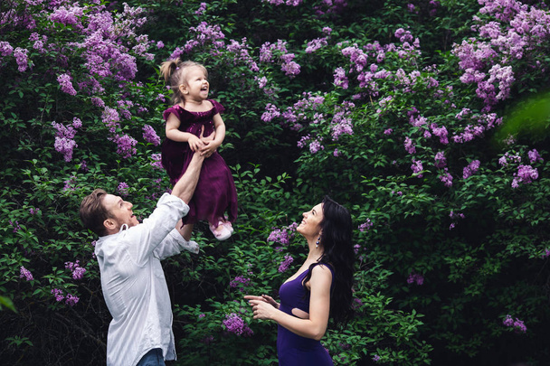 cheerful happy family near lilac flowering bushes. dad throws up little daughter up - Photo, Image