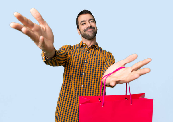 Mann mit Einkaufstaschen präsentiert und lädt ein, mit der Hand auf isoliertem blauem Hintergrund zu kommen - Foto, Bild