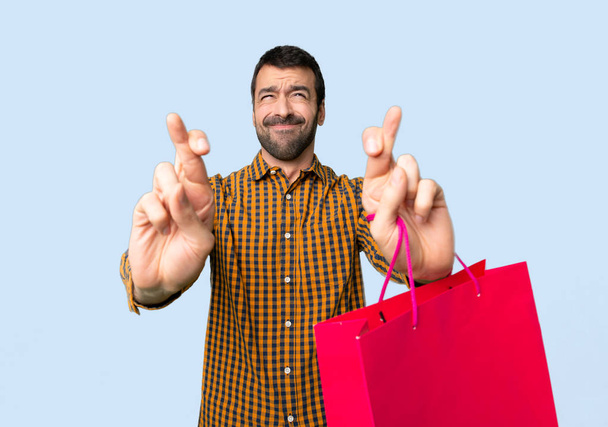 Man with shopping bags with fingers crossing and wishing the best on isolated blue background - Φωτογραφία, εικόνα