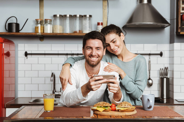 Happy young couple sitting at the kitchen during breakfast at home, using mobile phone - Zdjęcie, obraz
