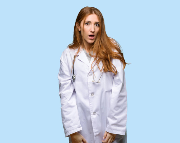 Pelirroja doctora con sorpresa y expresión facial impactada sobre fondo azul aislado
 - Foto, Imagen