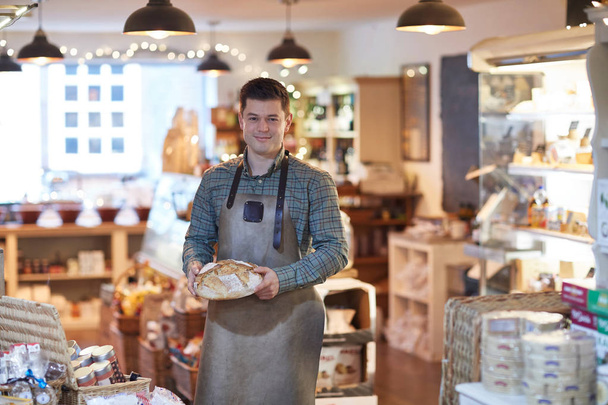 Portret uśmiechnięty mężczyzna właściciel sklepu Delikatesy na sobie fartuch trzyma bochenek chleba - Zdjęcie, obraz