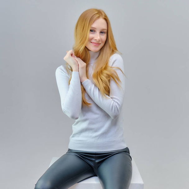 Concept portrait of a cute beautiful red-haired girl talking on a gray background in the studio. She is wearing a white sweater and shows different emotions in different poses. - Zdjęcie, obraz