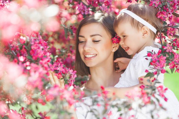 Muotokuva nuoresta kauniista äidistä pienen tyttövauvansa kanssa. Lähellä yhä rakastavaa perhettä. Houkutteleva nainen pitää lastaan vaaleanpunaisissa kukissa ja hymyilee
. - Valokuva, kuva