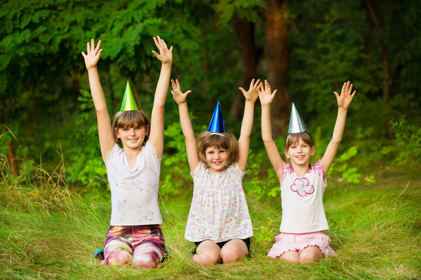 drei freundliche Kinder in festlichen Kegelmützen, sitzen auf dem Rasen, haben gemeinsam Spaß als Geburtstagskind mit fröhlichem Gesichtsausdruck in die Kamera, spielen gerne. - Foto, Bild