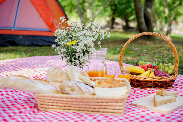 Cesta de mimbre de picnic con comida, pan, fruta y jugo de naranja sobre un paño rojo y blanco en el campo con fondo verde de la naturaleza. Concepto de picnic
. - Foto, imagen