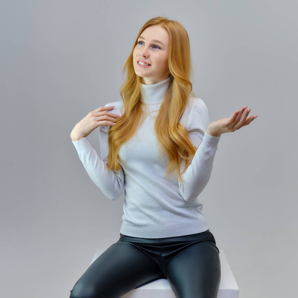 Concept portrait of a cute beautiful red-haired girl talking on a gray background in the studio. She is wearing a white sweater and shows different emotions in different poses. - Foto, Imagem