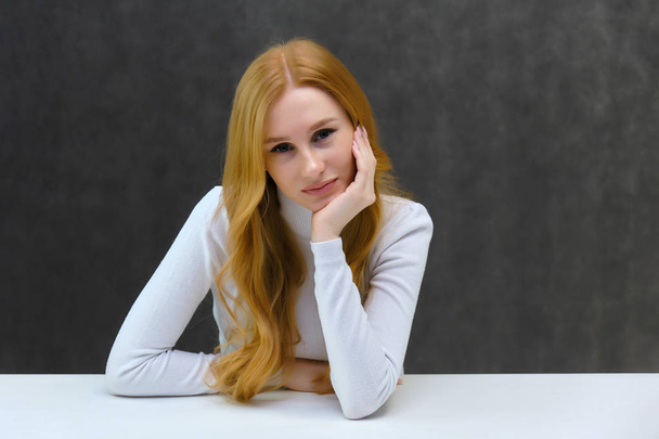 Concept portrait of a nice beautiful red-haired girl talking on a gray background in the studio sitting at the table. She is wearing a white sweater and shows different emotions in different poses. - Фото, зображення