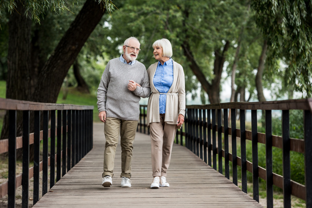 smiling senior couple talking while walking across wooden bridge in park - Photo, Image