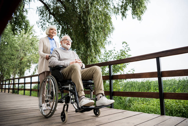 mujer mayor agradable llevando marido en silla de ruedas mientras camina en el parque
 - Foto, imagen