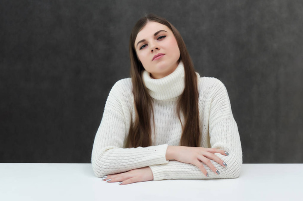 Concept portrait of a pretty beautiful brunette girl talking on a gray background in the studio sitting at the table. She is wearing a white sweater and shows different emotions in different poses. - Photo, image