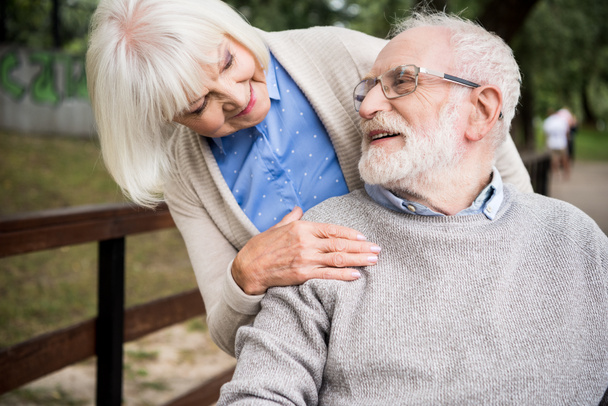 uśmiechający się starszy Kobieta, patrząc na szczęśliwy mąż na wózku inwalidzkim, trzymając rękę na jego ramieniu - Zdjęcie, obraz